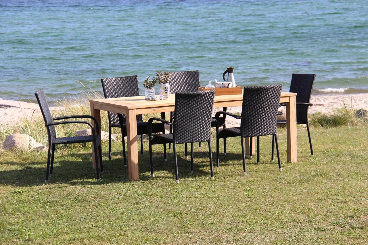 Udendørs teak spisebord med seks flettede havestole på græsplæne ved stranden